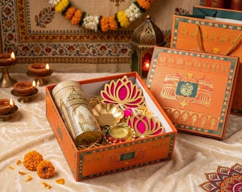 May include: An orange gift box containing a cylindrical tin with architectural illustrations, two pink lotus-shaped cutouts, a small gold-toned bowl, and a decorative candle holder with colorful beads, all nestled on shredded paper filler.