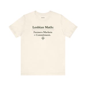 May include: A cream-colored short-sleeve t-shirt featuring the dark text "Lesbian Math: Farmers Markets = Commitment." with a small line drawing of a plant with a heart-shaped leaf at the bottom center and the brand name "StimChic" at the neck.