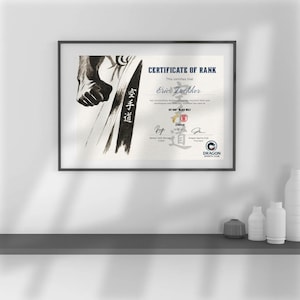 May include: Framed certificate of rank with the text "CERTIFICATE OF RANK" and "Eric Luckker". The certificate features a black and white illustration of a martial artist and the words "1ST DAN BLACK BELT". The Dragon Sports Club logo is visible.