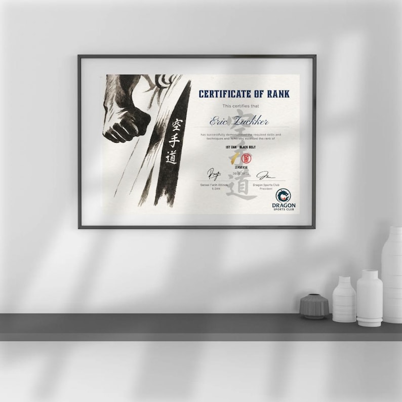 May include: Framed certificate of rank with the text "CERTIFICATE OF RANK" and "Eric Luckker". The certificate features a black and white illustration of a martial artist and the words "1ST DAN BLACK BELT". The Dragon Sports Club logo is visible.
