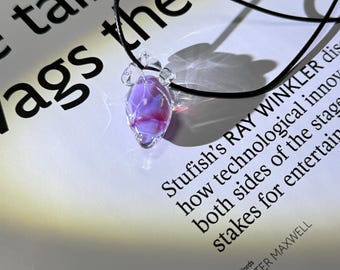 Puede incluir: Un colgante de cristal transparente con un diseño morado y rojo, suspendido de un cordón negro. El colgante es facetado y tiene forma de lágrima. El fondo incluye texto de un libro.