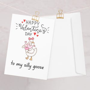Può includere: Un biglietto bianco di San Valentino con un'oca dei cartoni animati che tiene in mano dei palloncini rosa a forma di cuore. Il biglietto recita "Happy Valentine's Day" e "to my silly goose" in corsivo nero. Il biglietto è attaccato a una corda con una clip dorata.