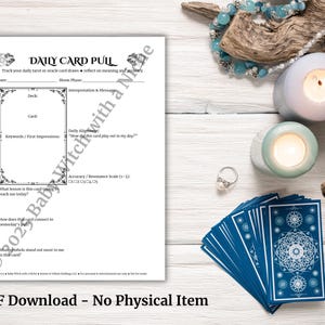 May include: A white printable daily card pull sheet with a decorative border, alongside a deck of blue tarot cards with white geometric designs. The image includes candles, jewelry, and a wooden accent.