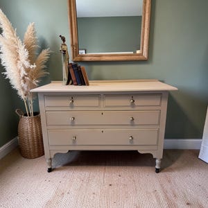 May include: A light beige chest of drawers with four drawers and brass knobs. A decorative mirror with a wooden frame hangs above the chest. A woven basket with tall, dried plants sits to the left. The room has a green wall and a woven rug.