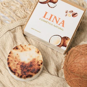 Könnte beinhalten: Ein rundes, handwerkliches Seifenstück mit hellbraunem, gesprenkeltem Design, neben einer Schachtel mit der Aufschrift "LINA Cosmética Natural". Die Schachtel zeigt Kokosnuss-Illustrationen. Eine ganze Kokosnuss und ein beiger Strickstoff vervollständigen die Szene.
