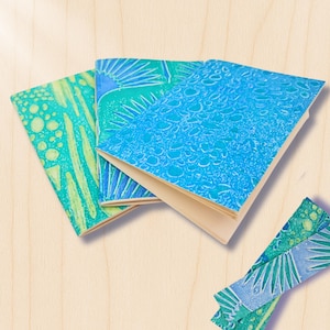 Può includere: Tre piccoli quaderni con copertine decorate nei toni del blu e del verde sono esposti su una superficie di legno chiaro. Le copertine presentano vari disegni, tra cui raggi di sole e forme organiche. Sono visibili anche alcune strisce decorative abbinate.
