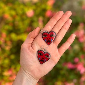 Puede incluir: Un par de pendientes en forma de corazón con base roja y contorno negro. Cada pendiente presenta una cara triste con lágrimas y pestañas. Los pendientes se sostienen en una mano sobre un fondo floral borroso.