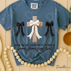 May include: A blue Comfort Colors t-shirt featuring a wooden cross with a white bow and two black bows. The text reads "NEW YEAR. NEW BEGINNINGS. Some Amazing God."
