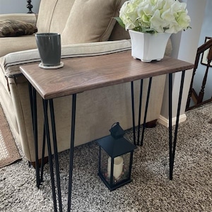 Op de afbeelding: Een kleine, rechthoekige houten bijzettafel met een donkere afwerking en zwarte haarspeldpoten. Op de tafel staat een witte vierkante vaas met witte bloemen en een grijze mok. Een zwarte lantaarn met een kaars staat op de vloer.