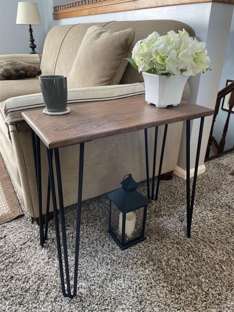May include: A small, rectangular wooden side table with a dark finish and black hairpin legs. A white square vase with white flowers sits on the table, along with a gray mug on a coaster. A black lantern with a candle is on the floor.