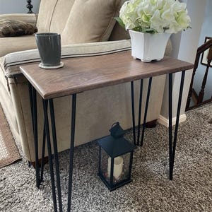May include: A small, rectangular wooden side table with a dark finish and black hairpin legs. A white square vase with white flowers sits on the table, along with a gray mug on a coaster. A black lantern with a candle is on the floor.