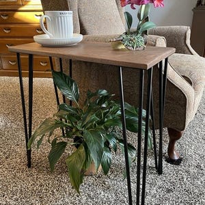 May include: A small, brown wooden side table with black hairpin legs. A white teacup and saucer with the text "ENJOY TODAY" sits on the table, along with a potted orchid. A potted plant is below the table.