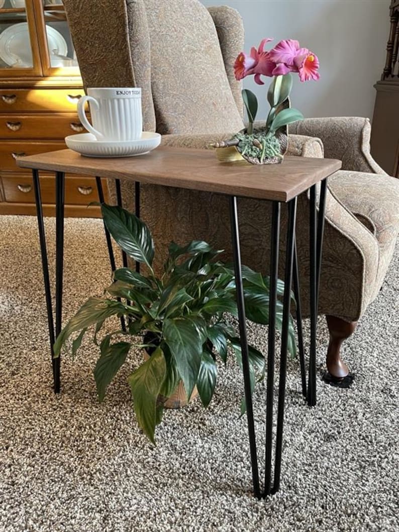 May include: A small, brown wooden side table with black hairpin legs. A white teacup and saucer with the text "ENJOY TODAY" sits on the table, along with a potted orchid. A potted plant is below the table.