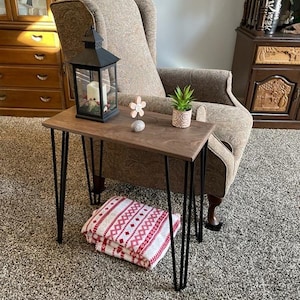 May include: A small, dark wood side table with black hairpin legs. A black lantern with a candle sits on the table, along with a small potted plant and decorative flower. A folded red and white patterned blanket rests on the floor.