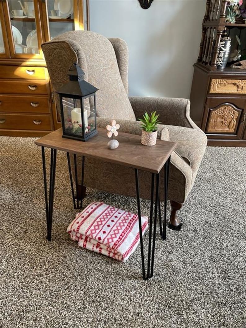 May include: A small, dark wood side table with black hairpin legs. A black lantern with a candle sits on the table, along with a small potted plant and decorative flower. A folded red and white patterned blanket rests on the floor.