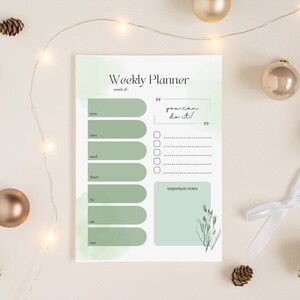 Op de afbeelding: Een weekplanner met een zachtgroen aquarelontwerp. De planner bevat secties voor elke dag van de week, een ruimte voor notities en de tekst "you can do it!". Decoratieve elementen zijn takjes bladeren en gouden ornamenten.