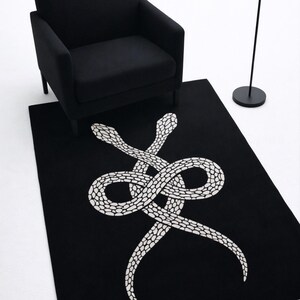 Può includere: Tappeto rettangolare nero con un disegno di serpente bianco. Il tappeto è posto su un pavimento bianco, con una poltrona nera e una lampada da terra nera sullo sfondo. Il disegno del serpente presenta un motivo a scaglie dettagliato.