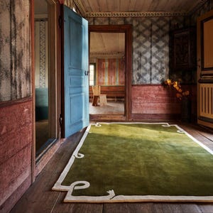 Op de afbeelding: Een groene rechthoekige vloerkleed met een witte rand en decoratieve hoekontwerpen ligt op een houten vloer. Het vloerkleed bevindt zich in een kamer met blauwe deuren en muren met een mix van kleuren en patronen.