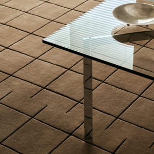 May include: A tan area rug featuring a grid pattern of recessed square lines. A glass-topped table with a metallic leg and a shallow silver bowl sits on the rug, which has a soft, textured surface and a consistent light brown color throughout.