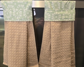 May include: Two kitchen towels hanging on an oven door. The towels are beige with a textured pattern. The top of each towel has a light green band with a white floral design. The oven display shows the time as 6:16.
