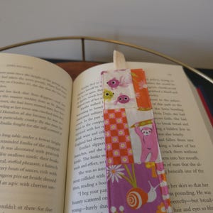 以下が含まれることがあります： パッチワークデザインの手作りファブリックブックマーク。ピンク、オレンジ、白の色合いのさまざまな模様の生地が特徴で、魚、カタツムリ、ナマケモノのイラストが描かれています。上部にはクリーム色のリボンが付いています。