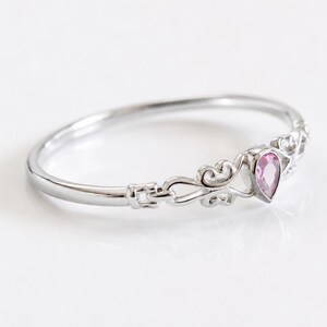 May include: A silver-toned ring featuring a pear-shaped pink gemstone set in a decorative metal frame. The band has a smooth, polished finish and connects to intricate, swirling metalwork that surrounds the central pink stone setting.
