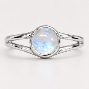 Pode incluir: Um anel de tom prateado com uma pedra da lua redonda de corte cabochão, apresentando um brilho azul translúcido e iridescente. A banda é desenhada com um estilo de haste dividida, onde o metal se separa em dois fios em direção ao engaste.