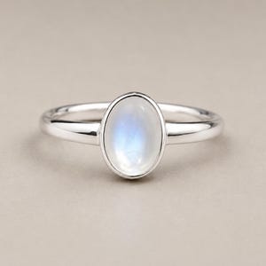 May include: A silver ring featuring a smooth, oval-shaped moonstone cabochon set in a polished metal bezel. The gemstone displays a translucent white appearance with a subtle blue adularescence, centred on a simple, rounded silver band.