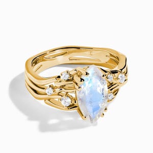 May include: A gold ring featuring a marquise-cut iridescent moonstone centre stone flanked by small round clear accent stones. The band has an organic, layered, and textured design with a 925 hallmark visible on the inner surface of the metal.