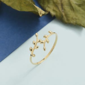 Op de afbeelding: Een gouden ring met een open bandontwerp, voorzien van twee delicate, symmetrische takjes versierd met kleine, gladde blaadjes. Het metaal heeft een gepolijste, reflecterende afwerking en de structuur is dun en licht, wat een doorlopend, vloeiend botanisch motief vormt dat gedeeltelijk om de vinger wikkelt.