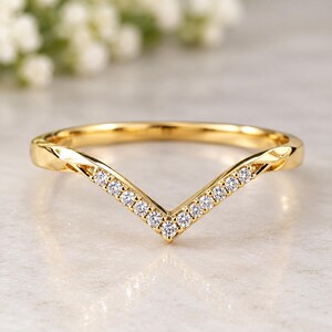 May include: A gold V-shaped chevron ring featuring a row of small, clear, round-cut stones set along the front band. The polished yellow gold band has a smooth, reflective finish and a pointed centre design that creates a distinct angular shape.