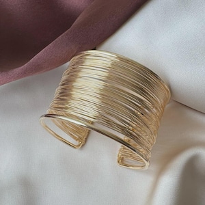 May include: A wide cuff bracelet constructed from multiple thin, polished gold-tone metal wires arranged in a parallel, layered design. The bracelet features an open-back style, allowing it to be slipped onto the wrist with ease.
