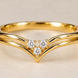 Op de afbeelding: Een gepolijste V-vormige ring van geelgoud met een gespleten schachtontwerp. Het midden van de chevronpunt is bezet met drie kleine, ronde, heldere edelstenen in briljantgeslepen vorm, gerangschikt in een driehoekige cluster op de band.