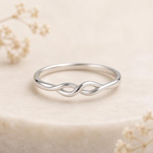 Peut inclure: Une bague en métal argenté poli présentant un motif central délicat en forme de nœud infini torsadé. L'anneau est lisse et fin, arborant une finition métallique continue qui se courbe pour former un motif de boucle symétrique sur le devant.