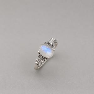 Könnte beinhalten: Ein Silberring mit einem ovalen Mondstein, der einen bläulichen, irisierenden Schimmer aufweist. Die Ringschiene ist auf beiden Seiten des Mittelsteins mit filigranen Metallarbeiten verziert, die ein fächerartiges Muster und feine Schnörkel zeigen.