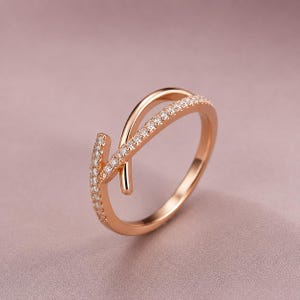 Könnte beinhalten: Ein Ring aus Roségold mit einem Bypass-Design aus zwei überlappenden Bändern. Ein Band ist glatt und poliert, während das andere mit einer Reihe kleiner, klarer, rund geschliffener Steine besetzt ist, die entlang der Vorderseite verlaufen.