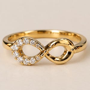 May include: A gold ring featuring an infinity symbol centrepiece. The left loop of the infinity design is set with ten small, clear, round-cut stones, while the right loop is crafted from polished, solid gold metal with a smooth, reflective surface.