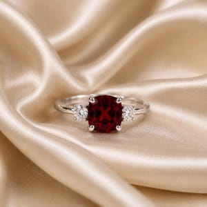 Puede incluir: Un anillo de tono plateado con una piedra preciosa central de color rojo intenso en talla cojín, flanqueada por dos piedras claras más pequeñas en talla redonda. La banda es de metal pulido y el engaste utiliza cuatro garras centrales.
