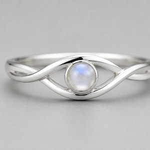 Könnte beinhalten: Ein Ring aus poliertem Silber mit einem zentralen, runden und schillernden Mondstein-Cabochon. Die Ringschiene besteht aus zwei glatten, ineinander verschlungenen Silbersträngen, die eine Zargenfassung um den Edelstein bilden.