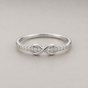 Può includere: Un anello color argento caratterizzato da un design centrale a forma di simbolo dell'infinito. La fascia è impreziosita da piccole pietre chiare a taglio rotondo incastonate lungo i lati, che conducono al motivo dell'infinito, creando un aspetto metallico lucido.