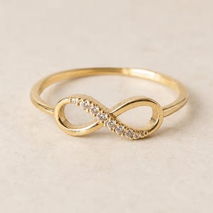 Peut inclure: Une bague en or avec un symbole de l'infini en pièce centrale. Un côté de la boucle de l'infini est orné d'une rangée de petites pierres claires et scintillantes, tandis que l'autre côté reste un anneau en or lisse et poli, fin et circulaire.