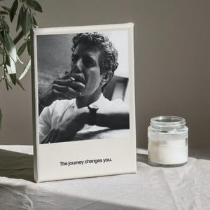 May include: Black and white canvas print featuring a portrait of a man smoking, with the text "The journey changes you." A small glass jar with a white substance sits to the right of the print. The print is on a neutral-colored surface.