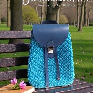 Könnte beinhalten: Ein türkisfarbener Häkelrucksack mit marineblauem Überschlag und Riemen, gesichert durch einen silbernen Verschluss. Die Tasche ist mit "Mini Lux" beschriftet und liegt auf einer Parkbank neben einem Buch und rosa Blumen. Der Text "Joconda's HM. Handcrafted Jewelry & Handbags" steht oben.