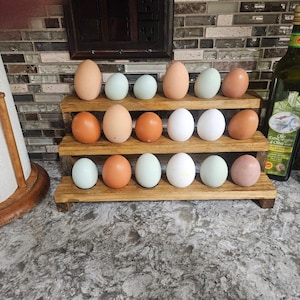 May include: A wooden tiered display holds a variety of eggs in shades of brown, blue, white, and pink. The eggs are arranged on three levels. A paper towel holder and olive oil bottle are visible in the background.