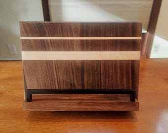 Puede incluir: Un atril de madera con acabado marrón oscuro, acentuado por una franja de color claro. Cuenta con un soporte de metal negro en la base, diseñado para sostener libros o tabletas. El atril es de madera y tiene vetas naturales.