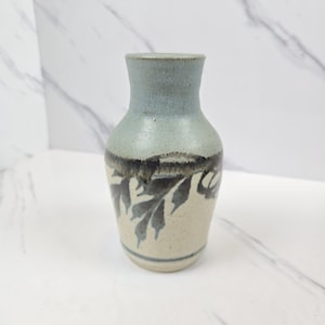 Puede incluir: Un pequeño jarrón de cerámica con la parte superior azul claro y la base beige. El jarrón presenta un diseño de hojas gris oscuro alrededor del centro. El jarrón mide aproximadamente 15 cm de altura y es adecuado para pequeños arreglos florales o decorativos.