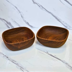 Könnte beinhalten: Zwei Holzschalen mit quadratischer Form und abgerundeten Ecken. Die Schalen sind aus dunkelbraunem Holz mit sichtbaren Maserungsmustern gefertigt. Die Schalen sind leer und stehen auf einer weißen Marmoroberfläche.