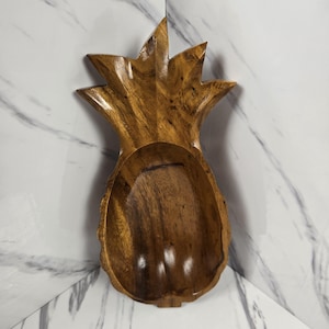 以下が含まれることがあります： 彫刻デザインの木製パイナップル型ボウル。ボウルは明るい茶色の木材でできており、深く丸みを帯びた内部があります。パイナップルの上部は尖った葉で詳細に描かれています。ボウルは、サービングまたはディスプレイ用に設計されています。