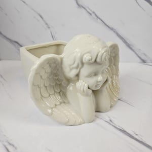 May include: Off-white ceramic planter in the shape of an angel. The angel is depicted with detailed wings and is resting its head on its hand. The planter has a rectangular opening for plants or decorative items. The overall design is smooth and elegant.
