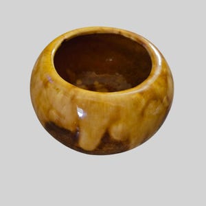 May include: A small, round ceramic bowl with a glossy finish. The exterior features a blended colour palette of warm honey yellow and deep brown, with a smooth, curved silhouette and a wide, open top that reveals a dark, reflective interior basin.
